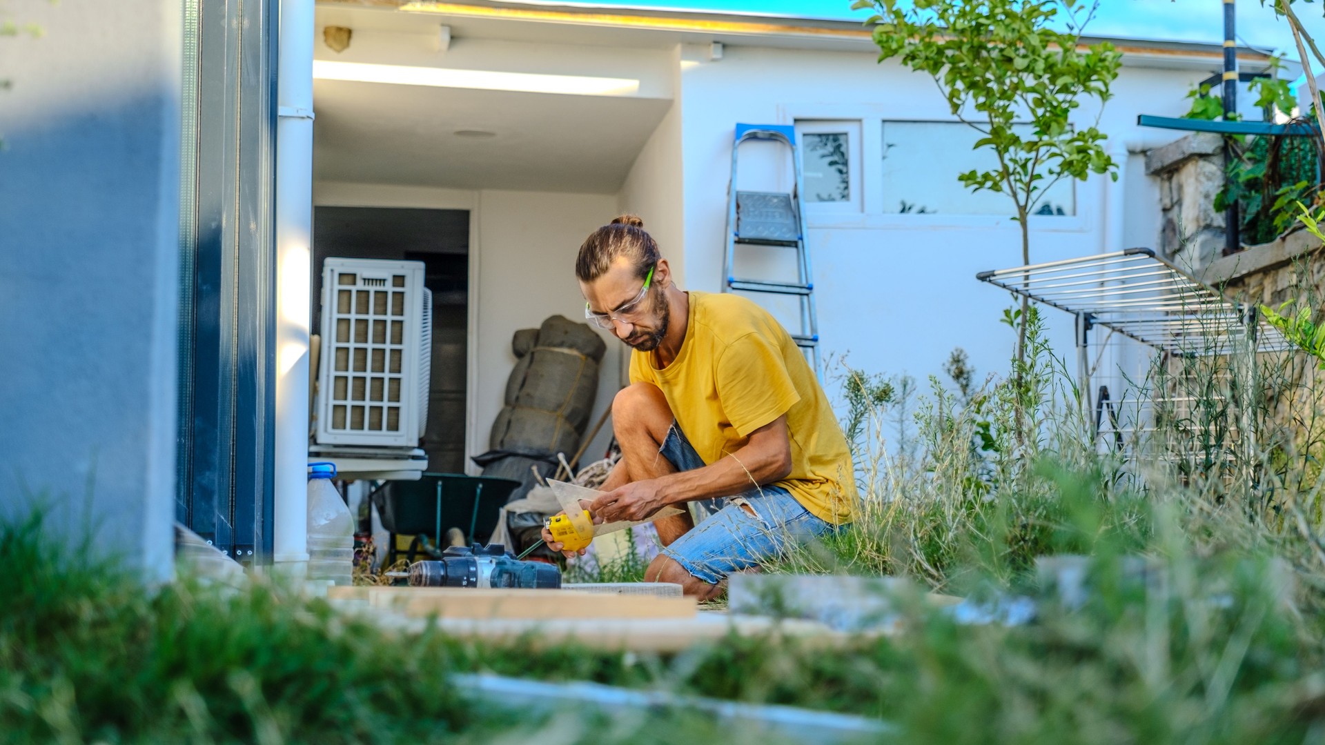 A middle-aged man measures with a tape measure to make repairs with lumber in his garden on a summer day